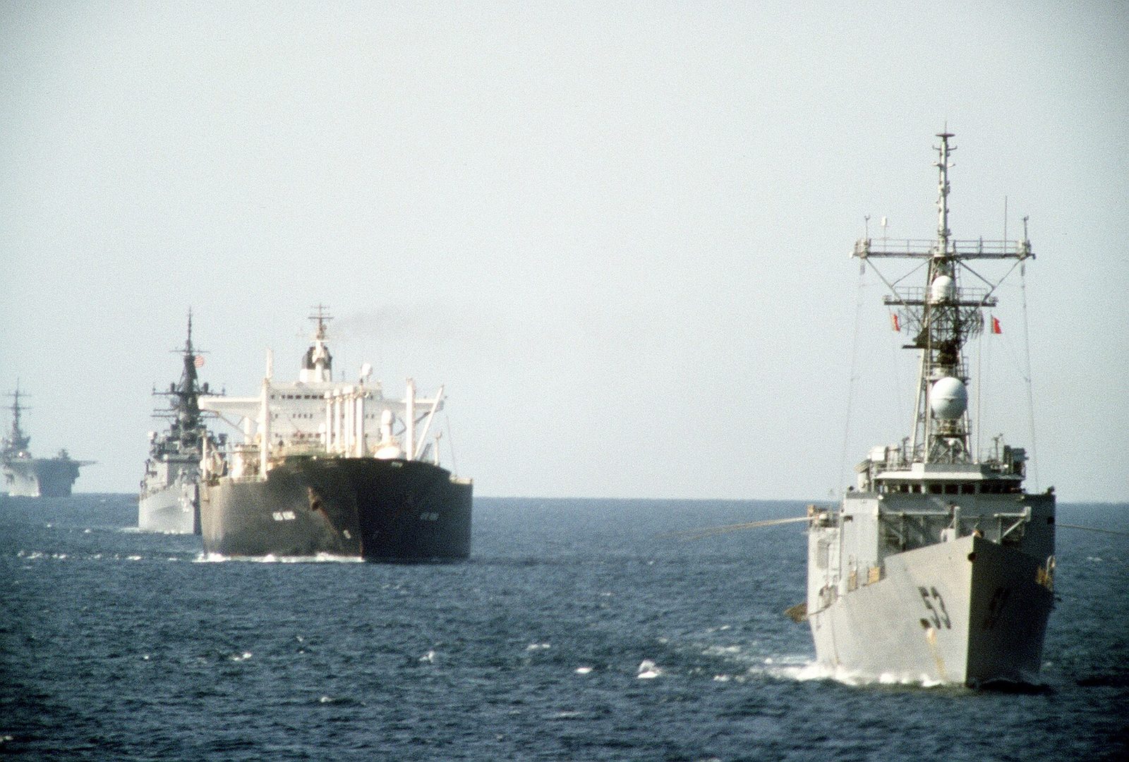 A 1987 photograph at sea: the frigate USS Hawes in the foreground, a large oil tanker astern of it with a cruiser and an amphibious ship following behind, all in a line on a calm grey sea