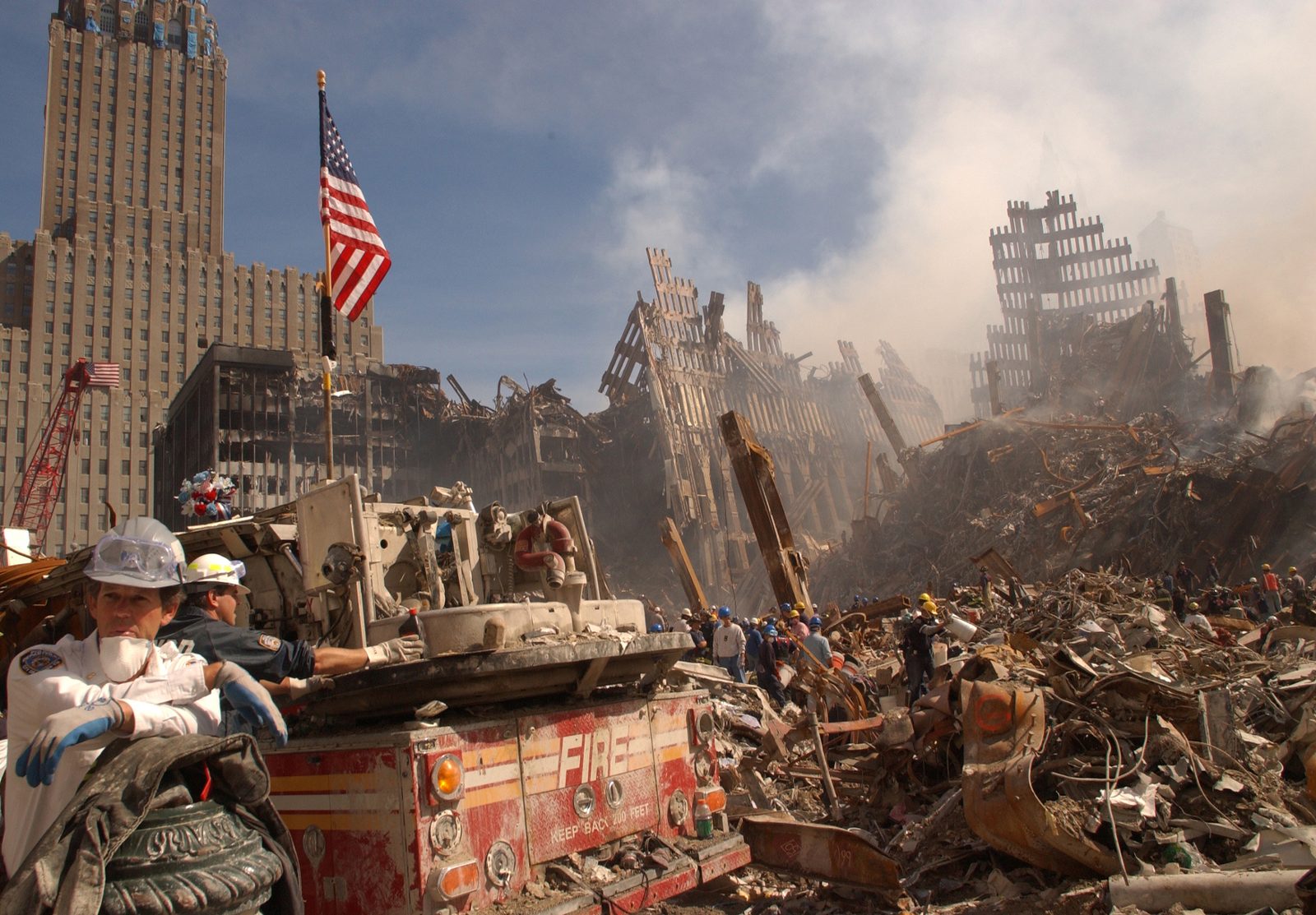 Ground Zero on 13 September 2001: rescue workers in dust masks and helmets in the foreground beside a crushed fire engine marked FIRE, an American flag on a pole above them, and behind them the shattered lattice of the towers' outer walls standing in the smoke and rubble