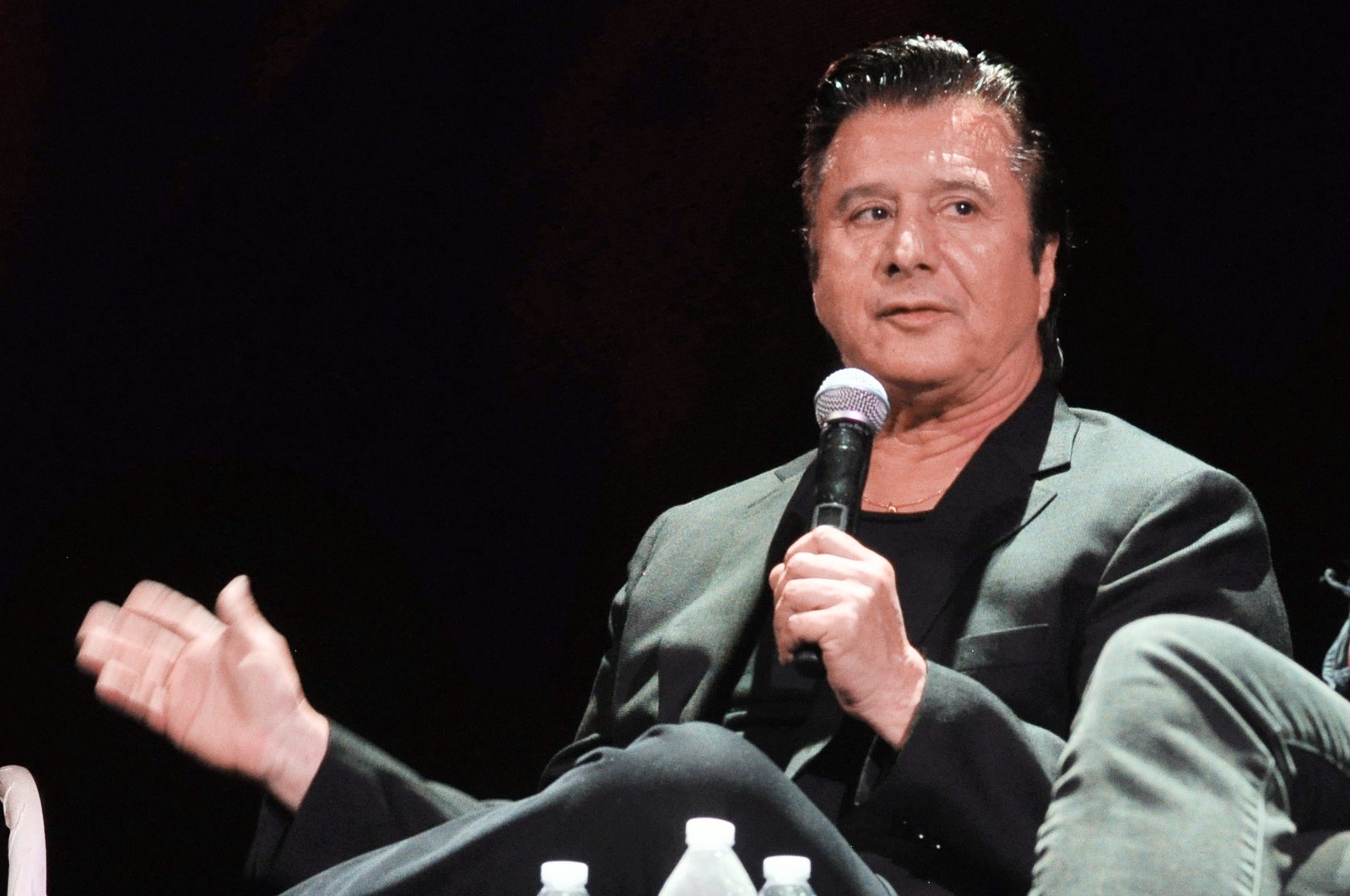 Steve Perry, older, with dark swept-back hair and a dark jacket over a black shirt, seated on a stage holding a microphone in one hand and gesturing with the other, mid-sentence, with a table of water bottles in front of him